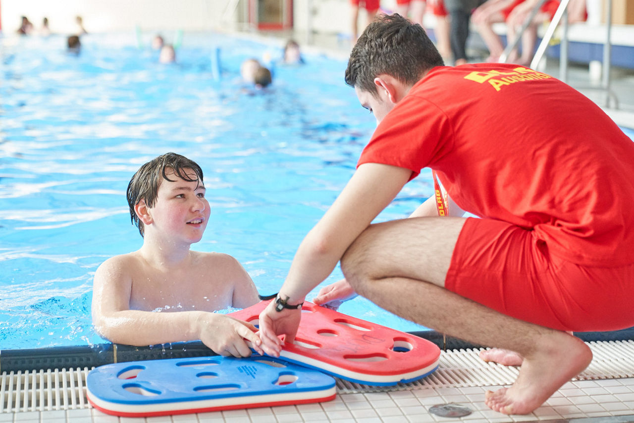 Ein Junge steht am Rand eines Wasserbeckens. Ein Schwimmlehrer reicht im eine Schwimmhilfe.