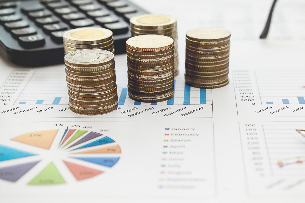 Gestapelte Euromünzen auf Finanzdokumenten mit Diagrammen, daneben ein Taschenrechner und ein Stift