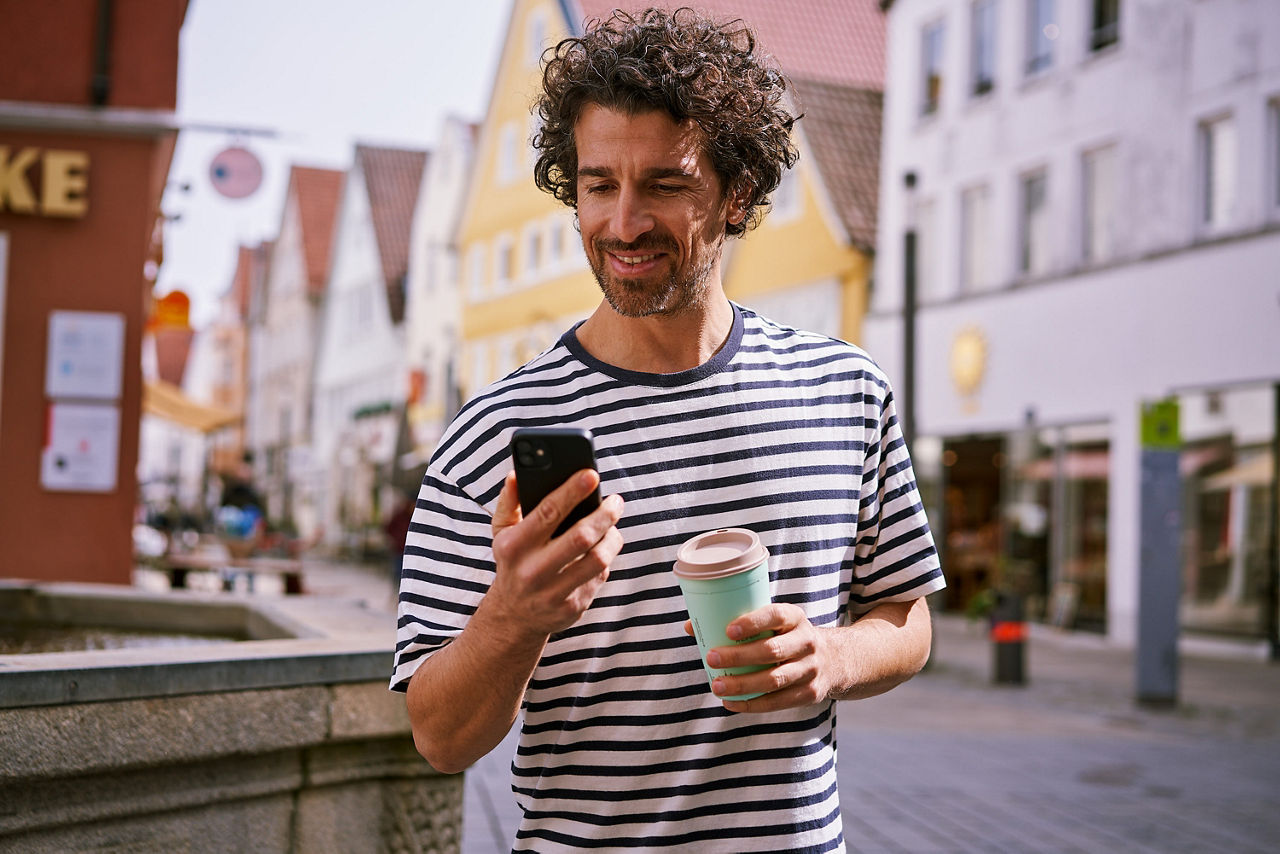 Mann mit Kaffeebecher steht in einer Einkaufsstraße und schaut auf sein Smartphone.
