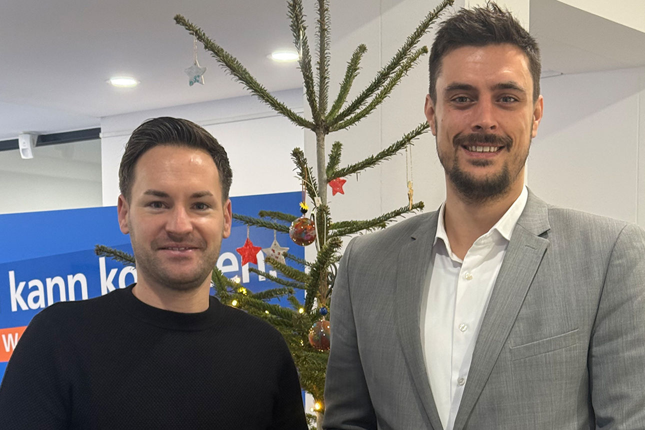 Zwei Männer lächeln in die Kamera. Sie halten einen Briefumschlag in der Hand.