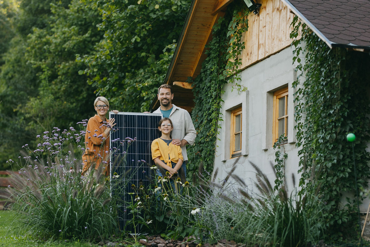 Dreiköpfige Familie steht im Garten vorm Haus mit Solar-Panel zwischen ihnen