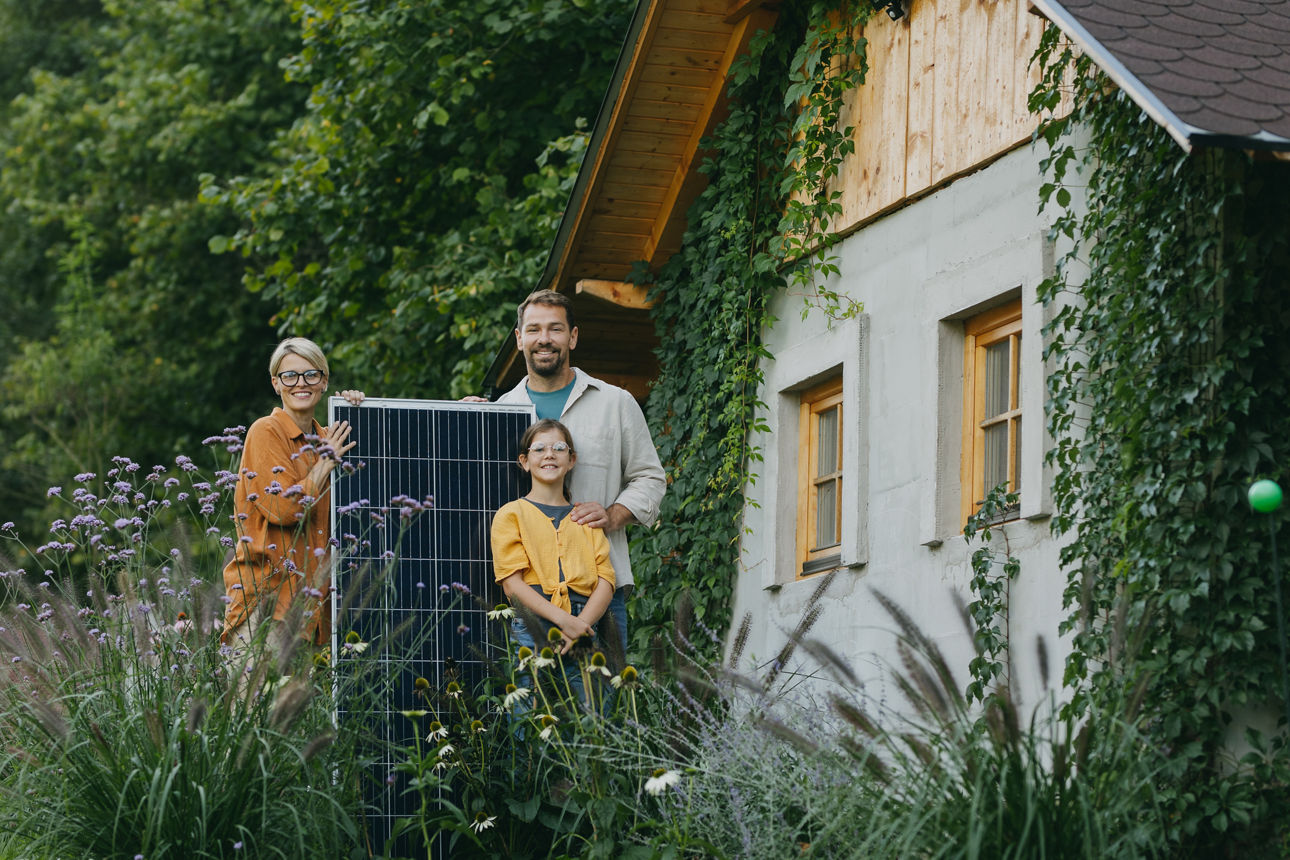 Dreiköpfige Familie steht im Garten vorm Haus mit Solar-Panel zwischen ihnen