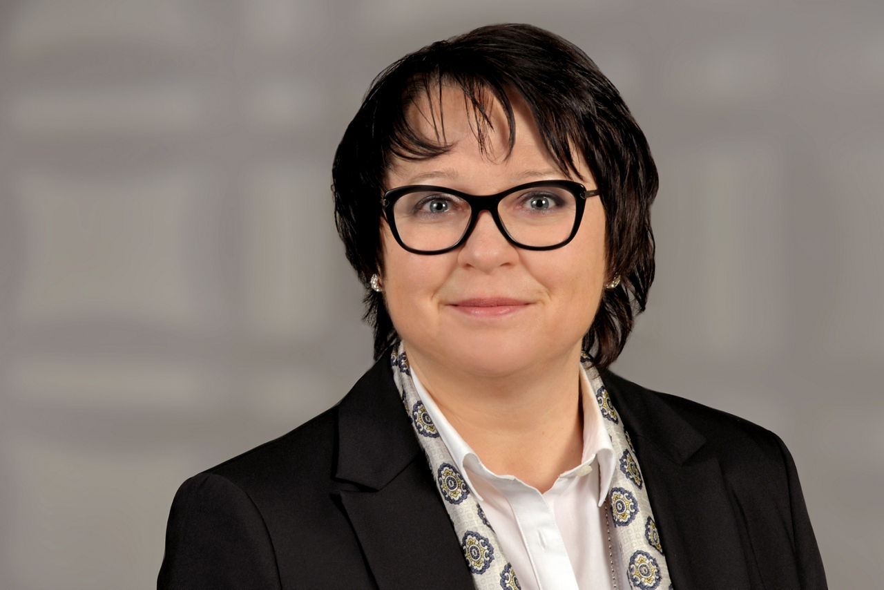 Frau mit dunklen Haaren und Brille trägt einen schwarzen Blazer, weißes Hemd und gemusterten Schal, steht vor neutralem Hintergrund