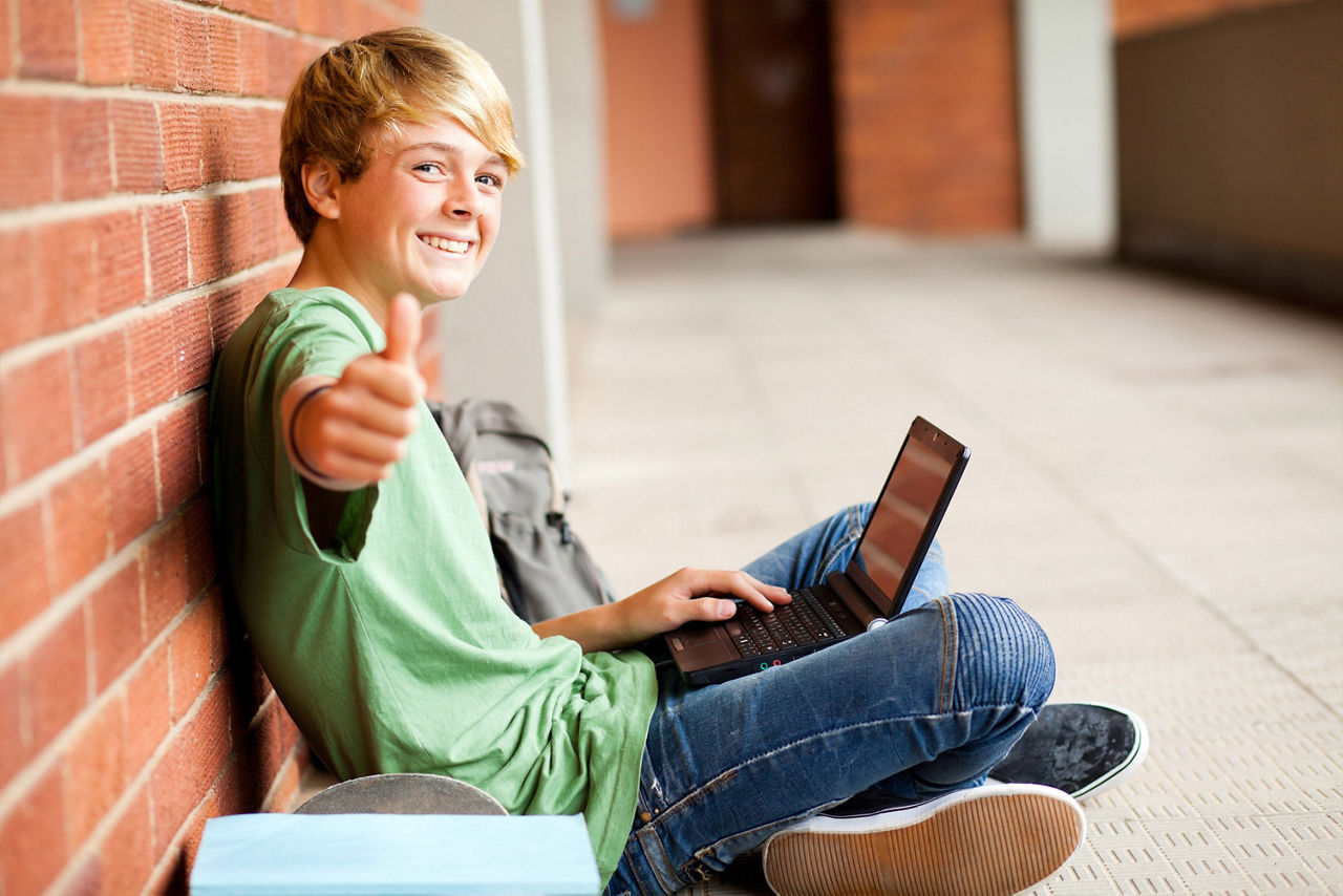 Schüler sitzt mit Laptop auf dem Schoß auf dem Schulflur und zeigt Daumen hoch in die Kamera