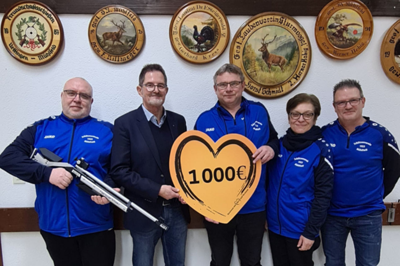 Fünf Personen stehen gemeinsam in einem Raum. Sie halten ein großes, herzförmiges Schild mit der Aufschrift 1000 Euro.