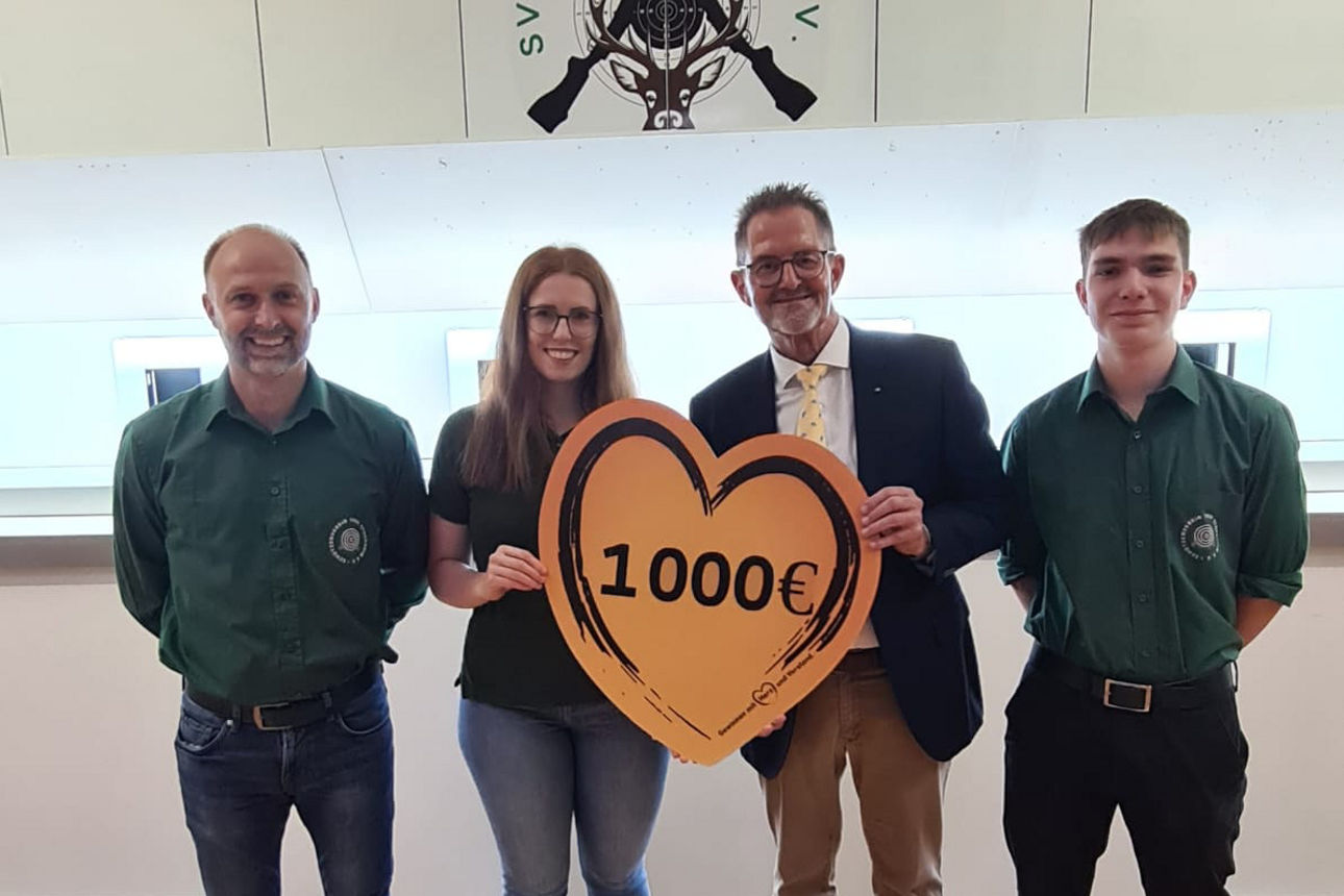 Vier Personen stehen nebeneinander und lächeln in die Kamera. Zwei von ihnen halten ein großes, orangefarbenes Herz mit der Aufschrift „1000 €“ in den Händen. Im Hintergrund ist das Logo des Schützenvereins „SV 1906 Stockheim e. V.“ zu sehen.
