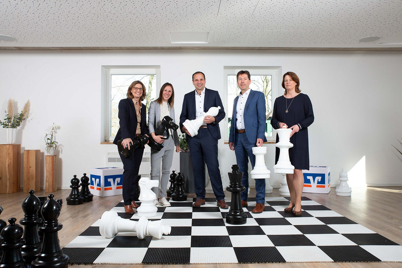 Unsere 5 Privat Banking Berater ( 3 Frauen, 2 Männer) stehen auf einem großen Schachbrett und halten große Schach Figuren in der Hand