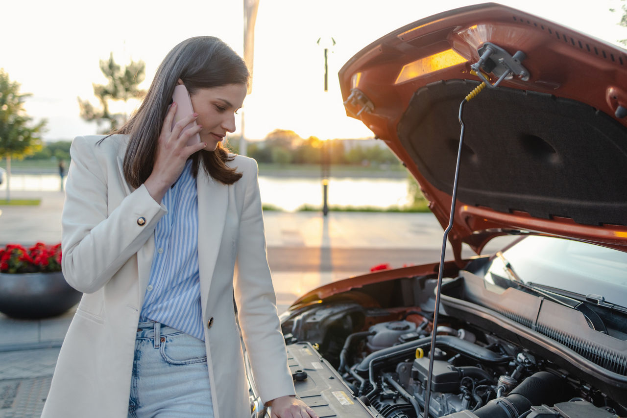 A young woman stands by her broken down car, making a phone call for roadside assistance. She appears calm and composed, set against a pleasant evening backdrop.