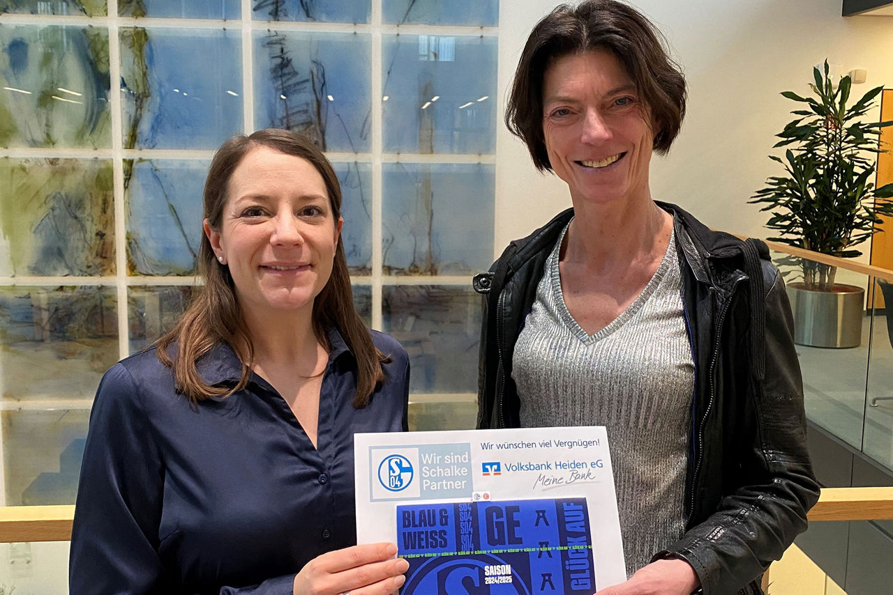 Zwei Frauen stehen vor einer Wand aus blauen und grünen Glasfliesen. Beide Frauen lächeln und halten eine weiße Urkunde mit e