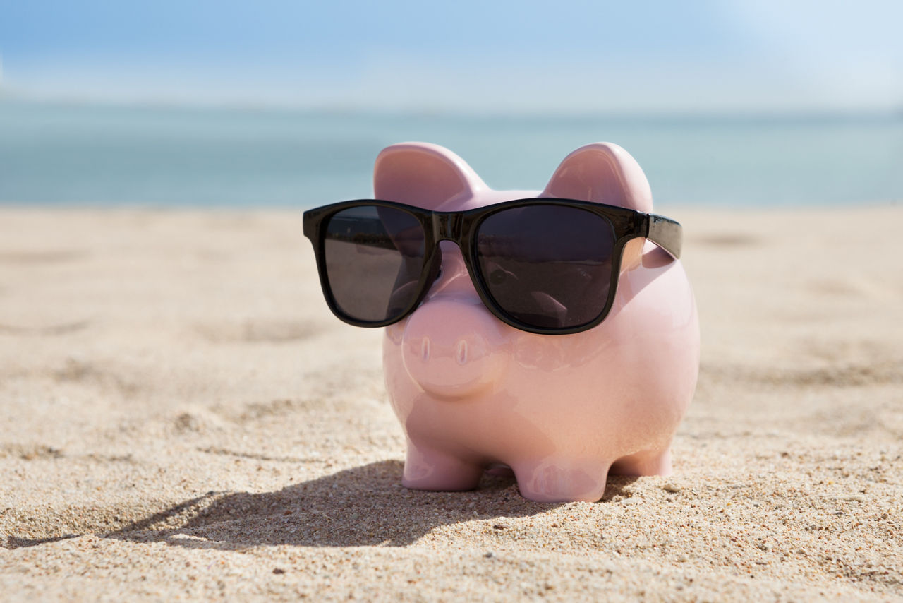 Ein rosa Sparschwein mit Sonnenbrille im Sand am Meer.