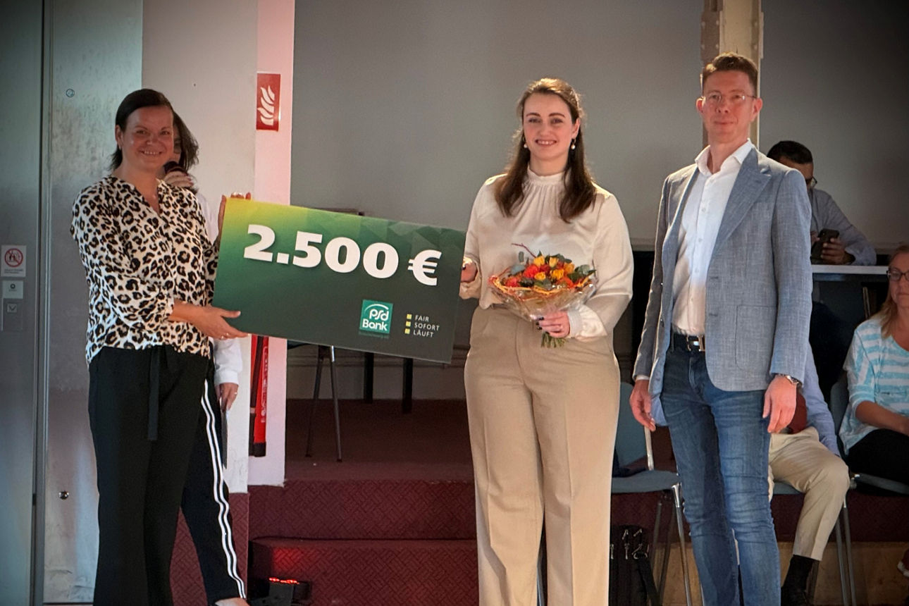 Eine Frau übergibt einem Mann und einer Frau einen Scheck in Höhe von 2.500 Euro