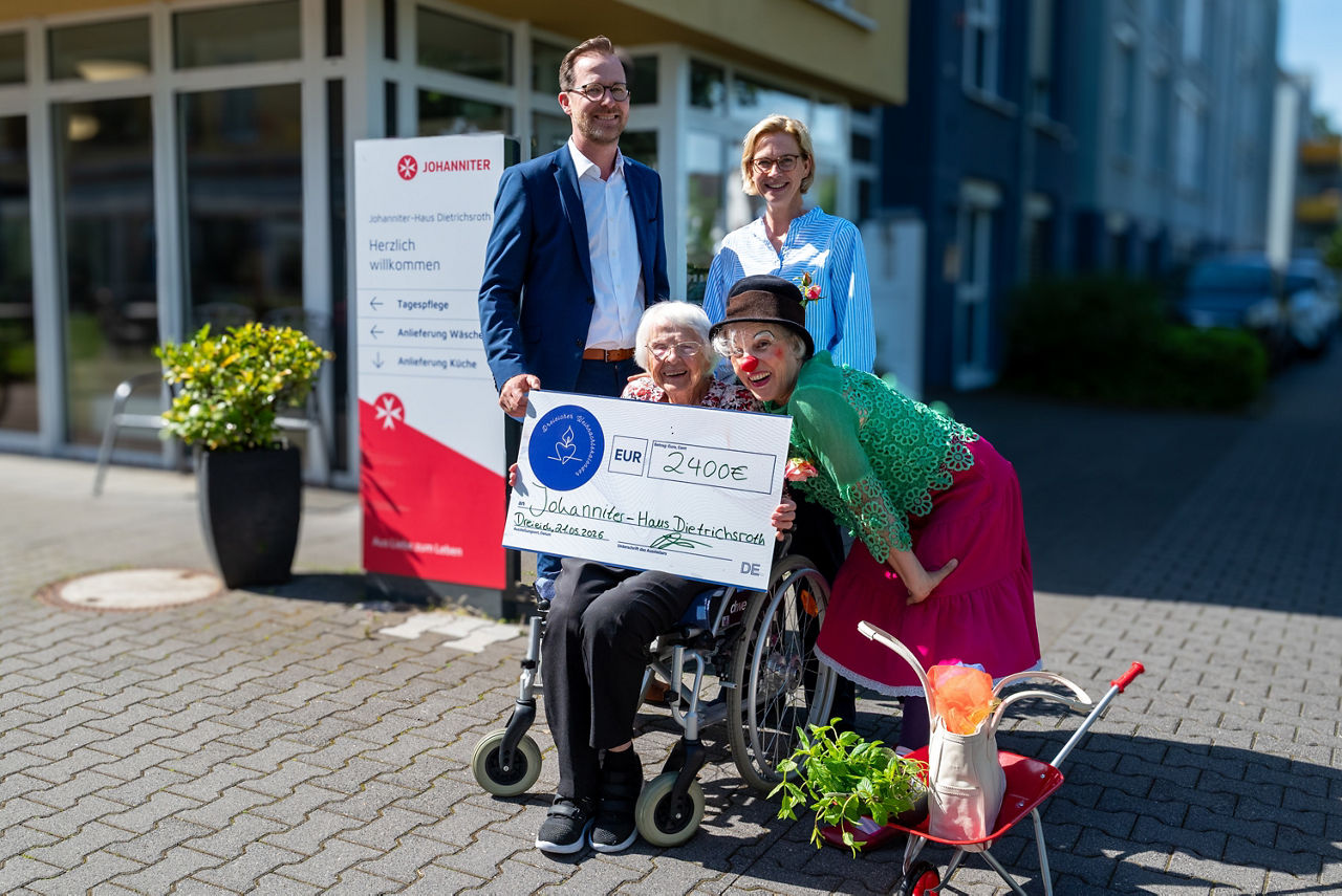 Mann im Anzug übergibt einer Clownin, einer Frau und einer älteren Dame im Rollstuhl einen Scheck über 2400€