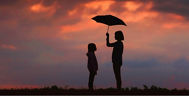 Frau und Kind stehen mit einem geöffneten Regenschirm vor einem bewölktem Himmel im Sonnenuntergang