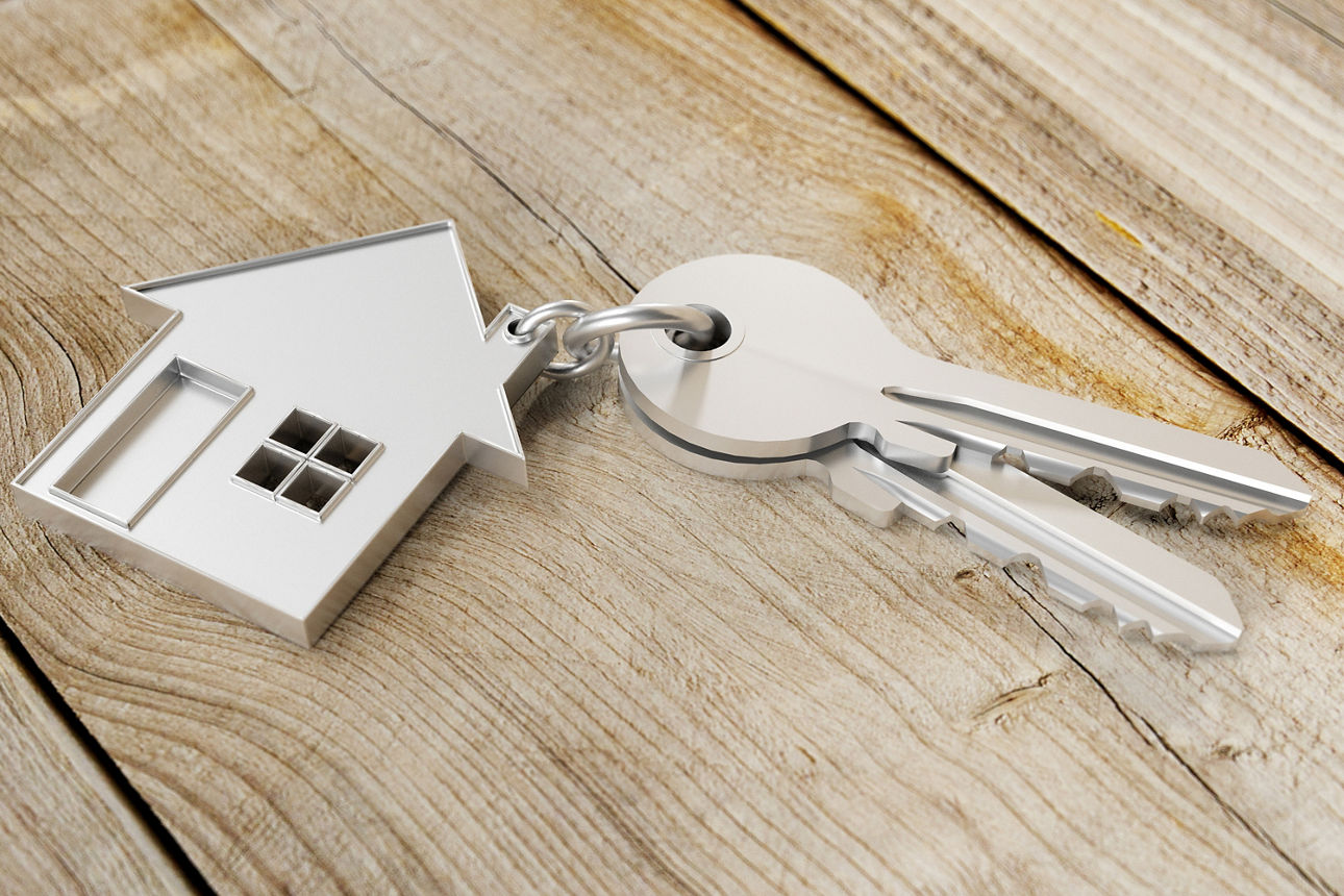 Auf einem Holztisch liegt ein silberner Schlüsselanhänger in Form eines Hauses. An dem Schlüsselanhänger befinden sich zwei Schlüssel.
