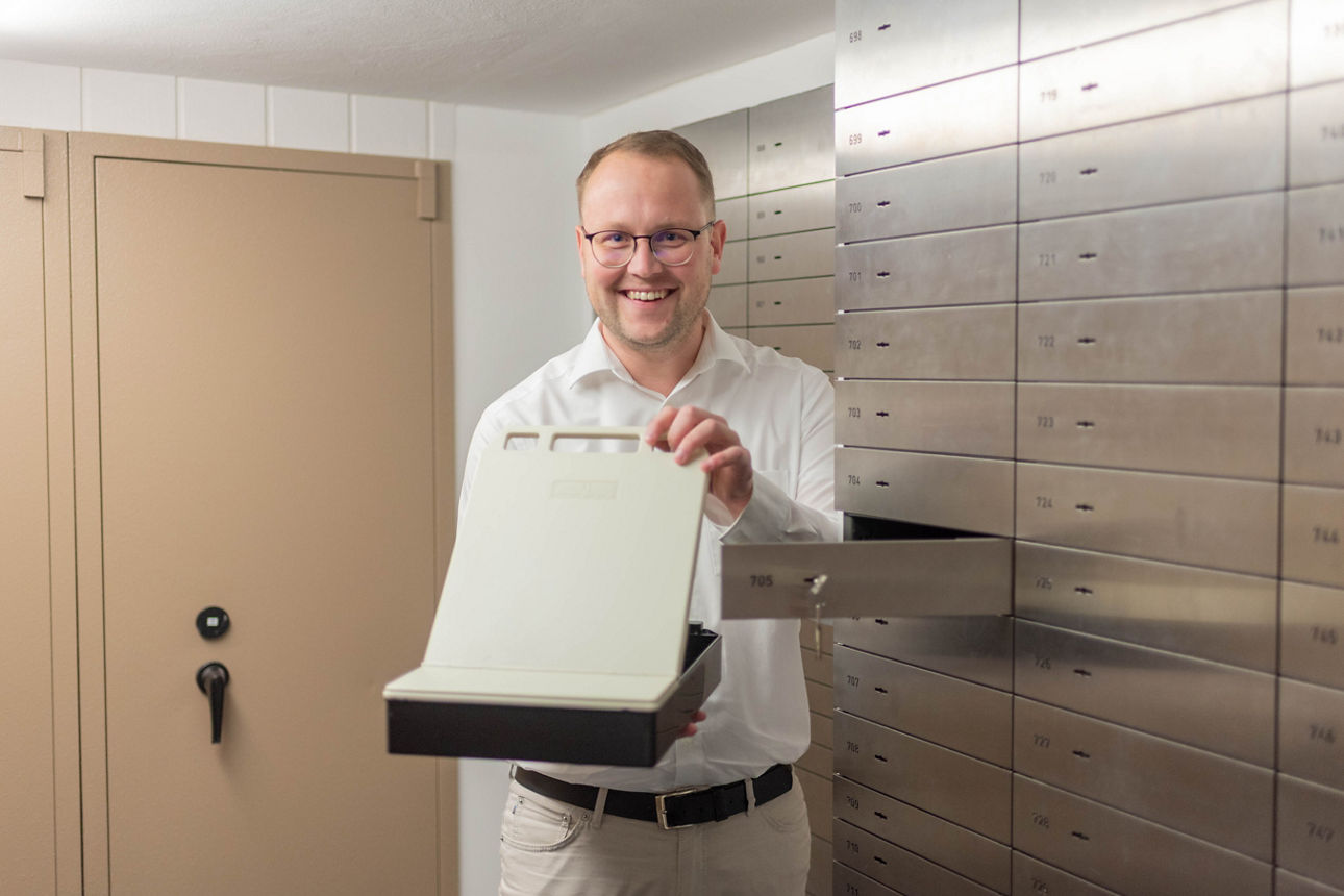 Junger Mann steht in einem Tresorraum vor einem Schließfach und hält die Schließfachkassette in der Hand.