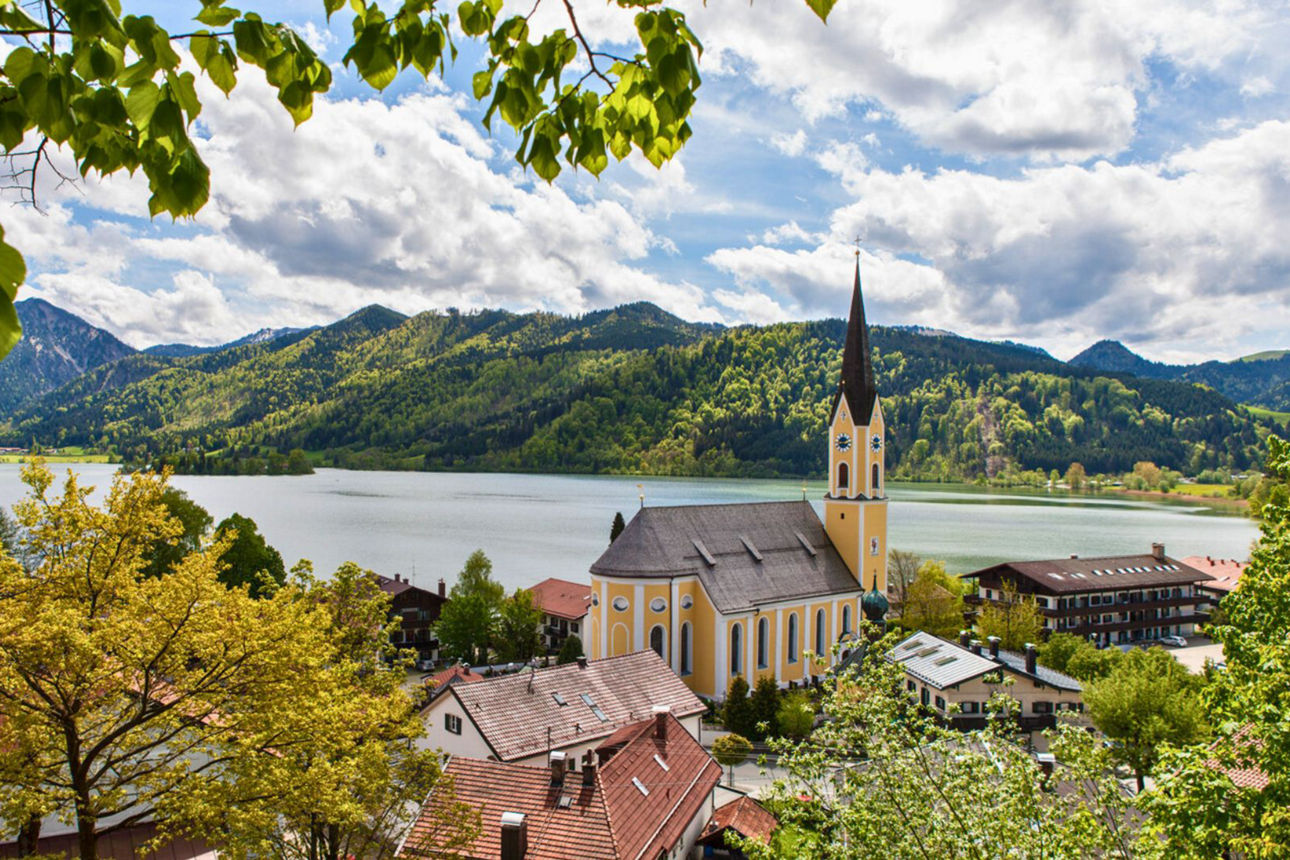 Eine malerische gelbe Kirche mit hohem, dunklem Zwiebelturm steht am Ufer des Schliersees, umgeben von Häusern und grünen Bäumen. Im Hintergrund erheben sich bewaldete Berge unter einem teilweise bewölkten Himmel.