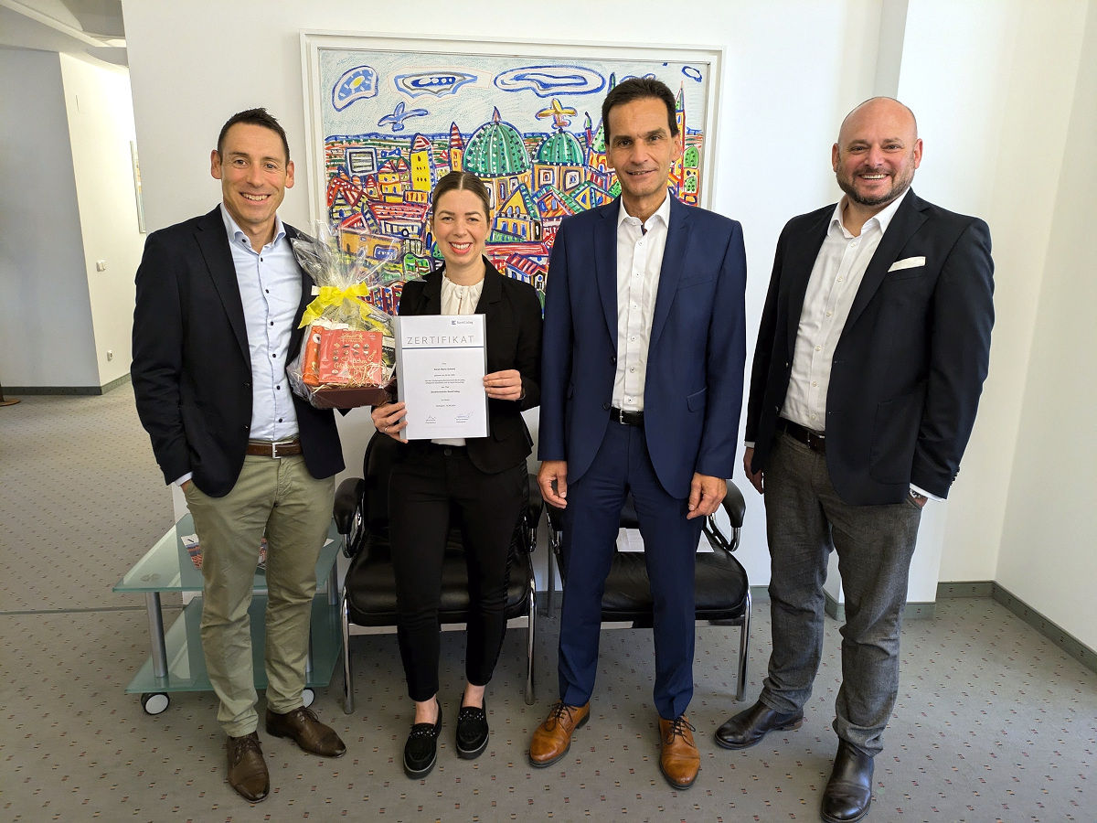 Mitarbeiterin steht mit Urkunde und Geschenkkorb neben drei Führungskräften vor einem Großen Bild
