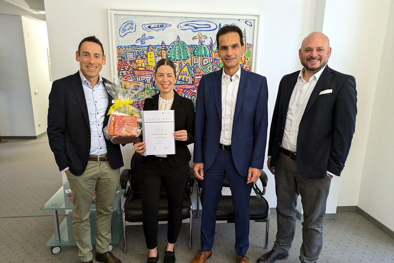 Mitarbeiterin steht mit Urkunde und Geschenkkorb neben drei Führungskräften vor einem Großen Bild