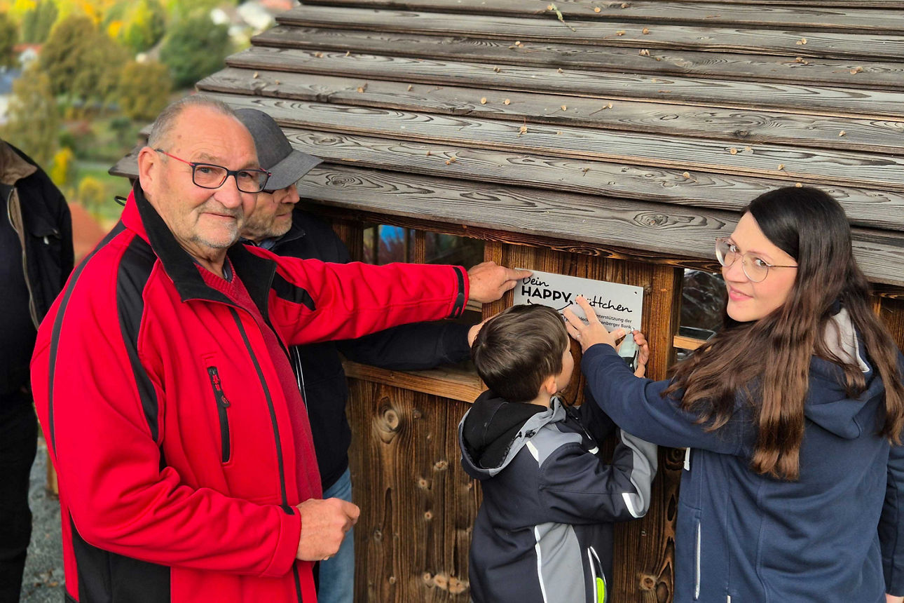 Mehrere Personen befestigen gemeinsam ein Schild mit der Aufschrift „Dein HAPPY, Plätzchen“ an einer hölzernen Schutzhütte.