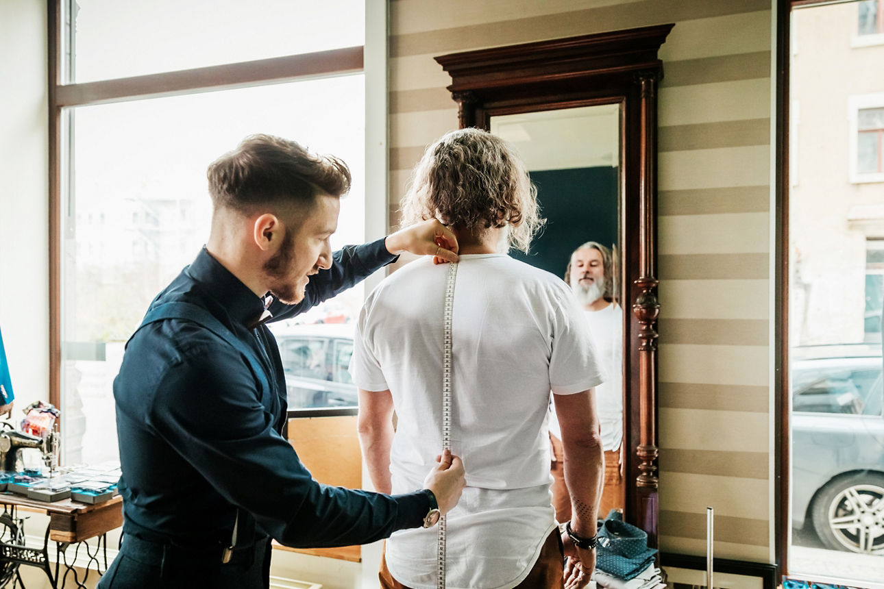 Schneider nimmt die Maße bei einem Mann auf, welcher mit weißem Hemd vor einem Spiegel steht