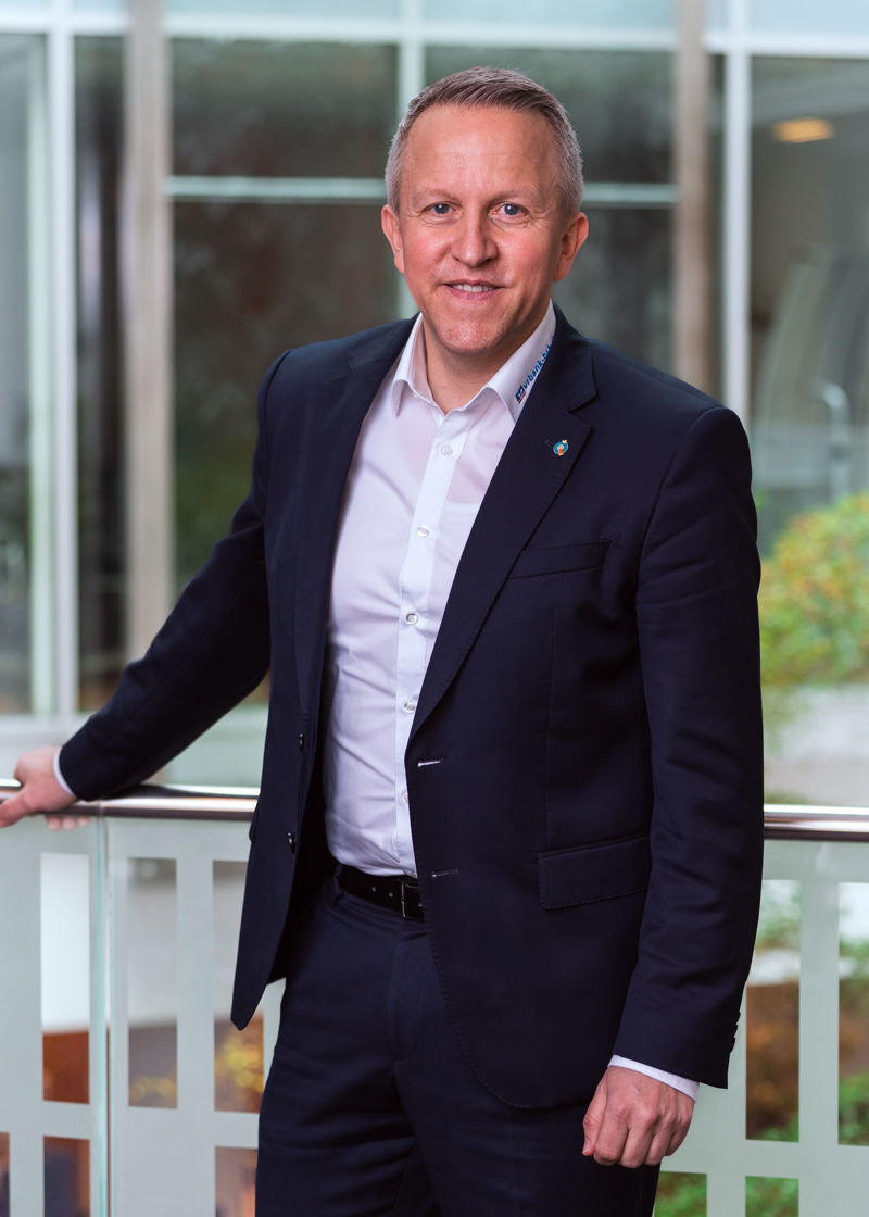 Ein Mann steht im dunklen Anzug vor einer Glaswand und lächelt in die Kamera.