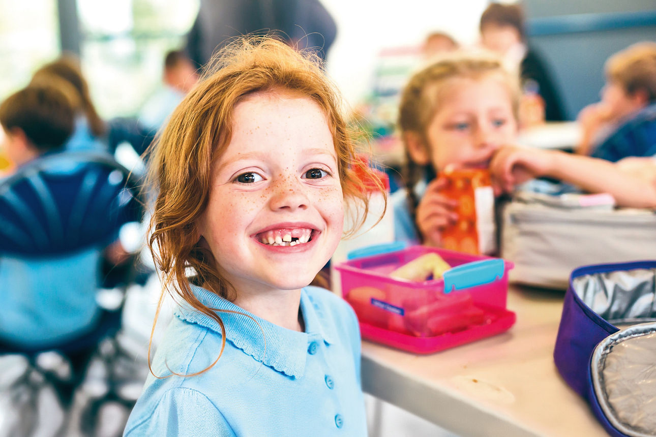 Lächelndes Mädchen am Tisch während der Schulpause mit weiteren Kindern im Hintergrund.