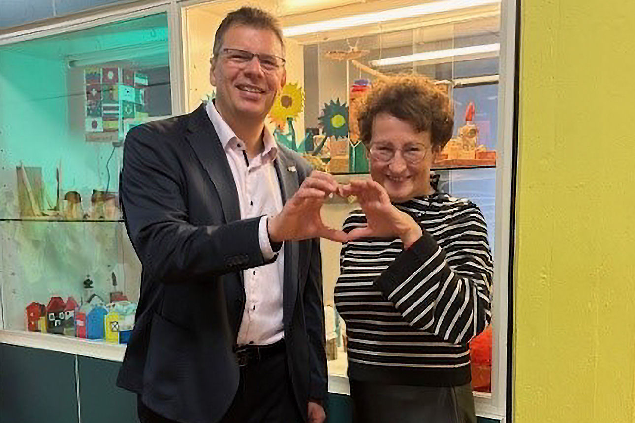 Ein Mann und eine Frau lächeln in die Kamera. Sie formen ein Herz aus ihren Händen.