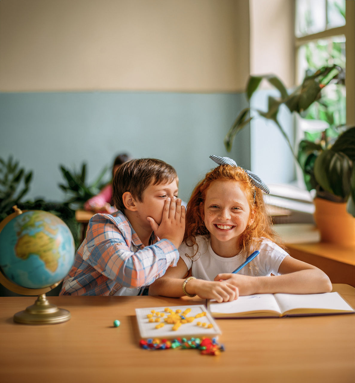 Zwei Kinder sitzen in einem Schulklassenraum an einem Tisch. Der Junge flüstert dem Mädchen etwas ins Ohr.
