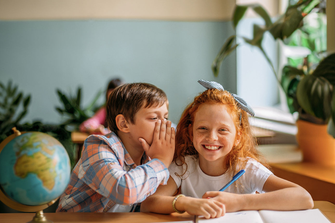 Zwei Kinder sitzen in einem Schulklassenraum an einem Tisch. Der Junge flüstert dem Mädchen etwas ins Ohr.
