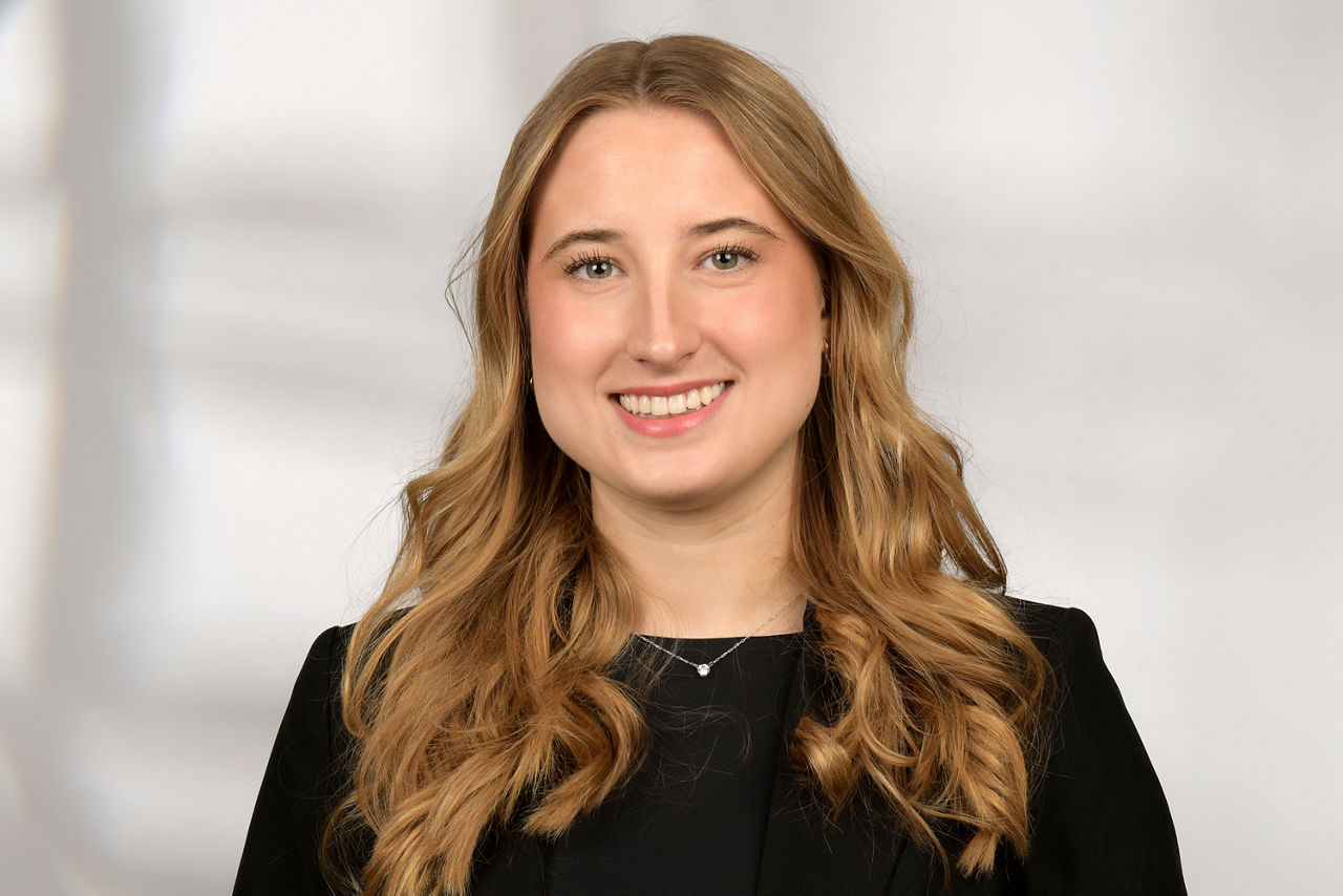 Frau mit langen, welligen Haaren trägt schwarzen Blazer und silberne Halskette vor unscharfem Hintergrund