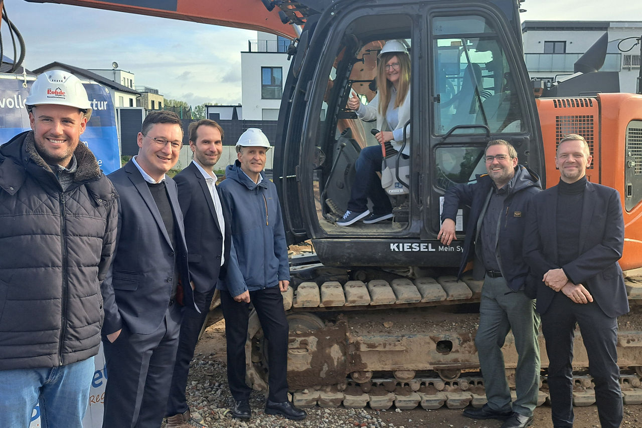 Sechs Männer stehen um einen Bagger herum. Im Bagger sitzt eine Frau mit Helm auf und lächelt. Sie befinden sich auf einer Baustelle.