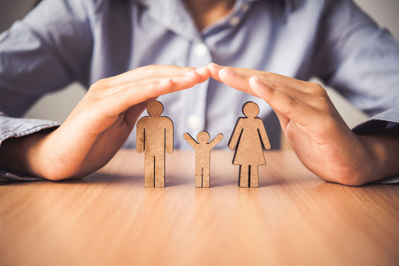 Zwei Hande bilden ein Dach über Holzfiguren die Familie darstellen, auf einem Tisch stehen