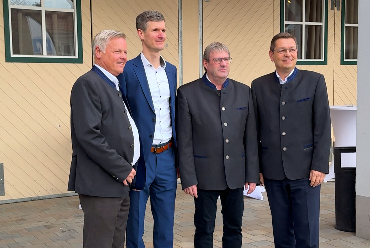 Vier Männer, drei in dunkelgrauem Janker, einer in blauem Anzug, stehen vor Gebäude und blicken  lächelnd zu einem imaginären Fotografen, der nicht im Bild zu sehen ist
