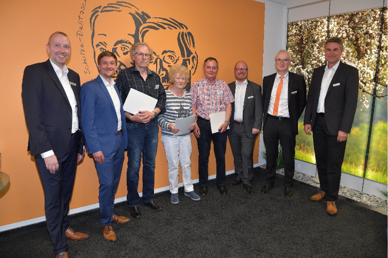 Eine Gruppe von sieben Personen steht vor einer orangefarbenen Wand und posiert für ein Foto.
