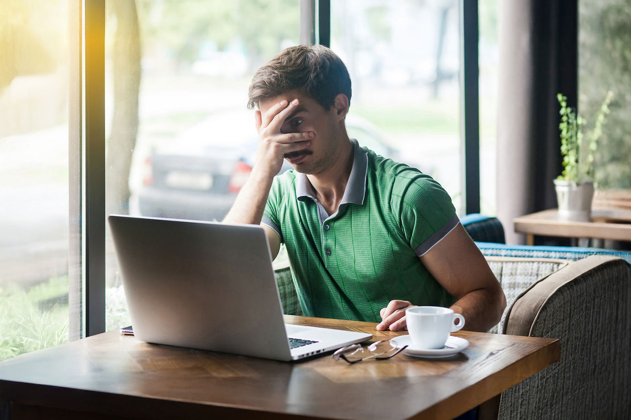 Mann schaut Schlechtes erwartend durch Hand hervor auf seinen Laptop