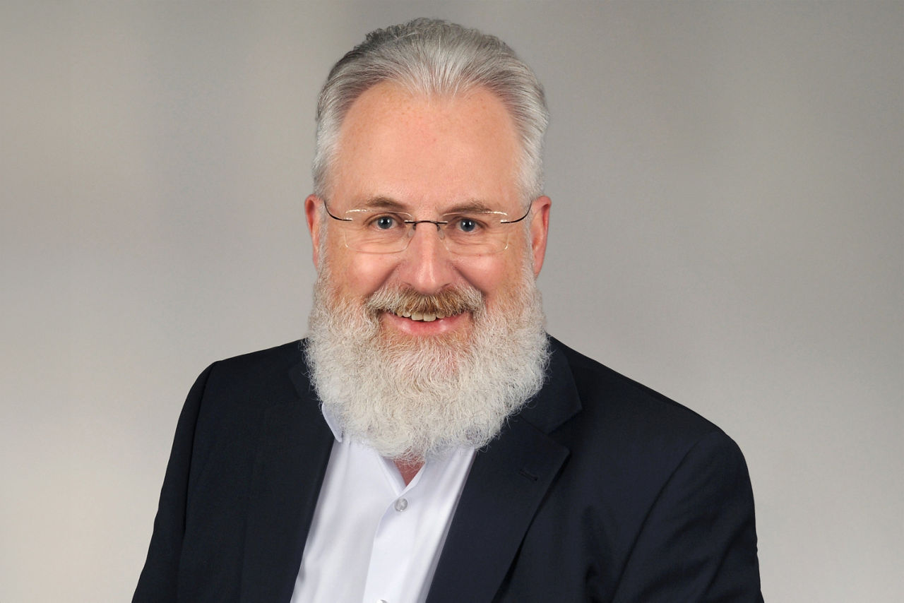 Älterer Mann mit grauem, zurückgekämmtem Haar und vollem weißem Bart trägt weißen Hemdkragen und schwarzen Anzug vor neutralem Hintergrund