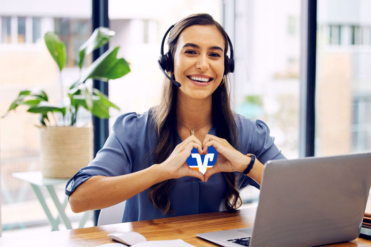 Frau sitzt mit Headset am Schreibtisch vor einem Laptop und hat die Hände zu einem Herz geformt, in dem das Markenzeichen der Volksbanken Raiffeisenbanken abgebildet ist