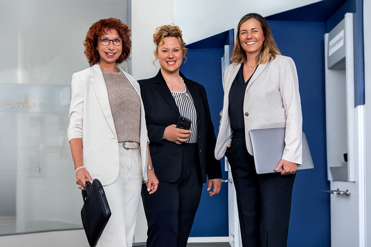 Drei Frauen in Businesskleidung vor einer blauen Wand mit Geldautomat in moderner Büro- oder Bankumgebung.