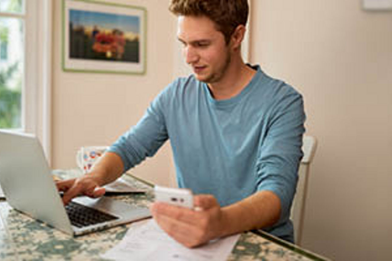 Junger Mann sitzt am Laptop und hält ein Smartphone in der Hand