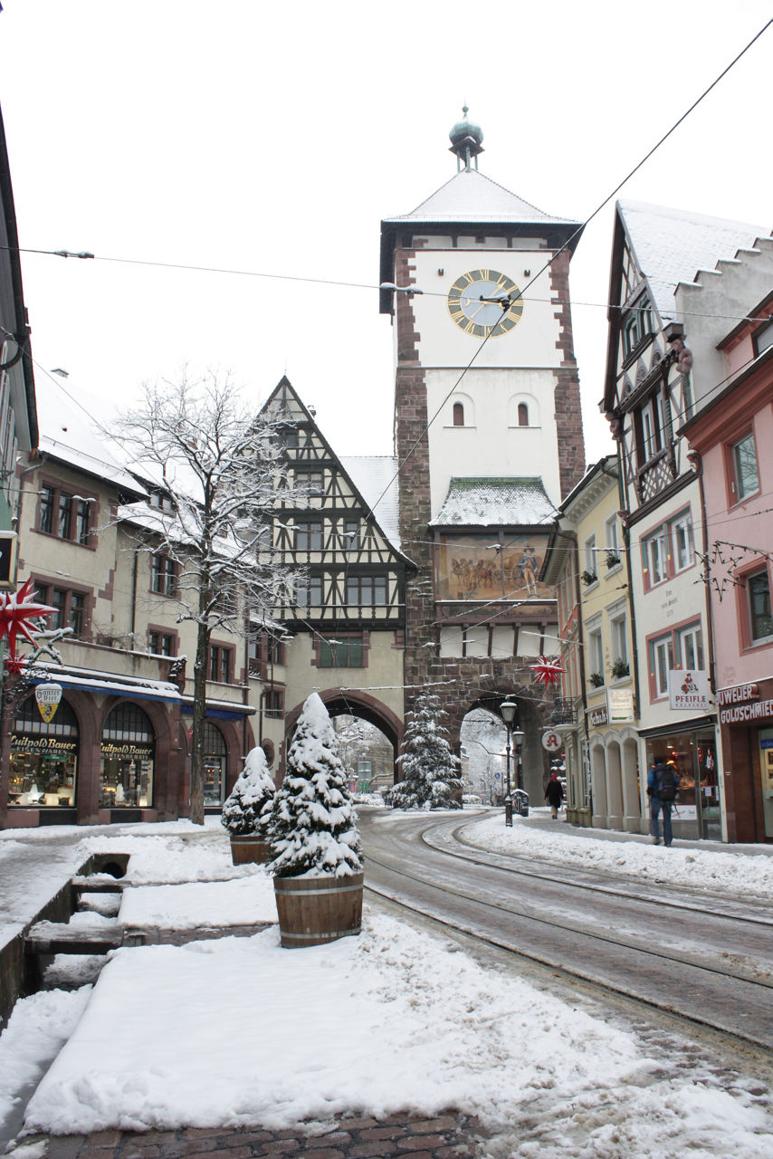 Das Bild zeigt ein verschneites Altstadt-Straßenbild mit einem historischen Stadttor samt Uhrturm in der Mitte, umgeben von Fachwerk- und Altbauten sowie weihnachtlich dekorierten Geschäften.