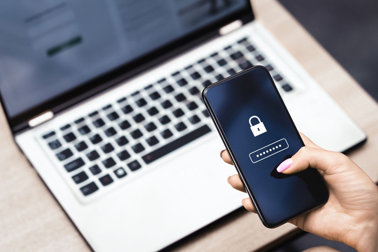 Frau hält Smartphone in der Hand. Im Hintergrund steht ein Laptop.