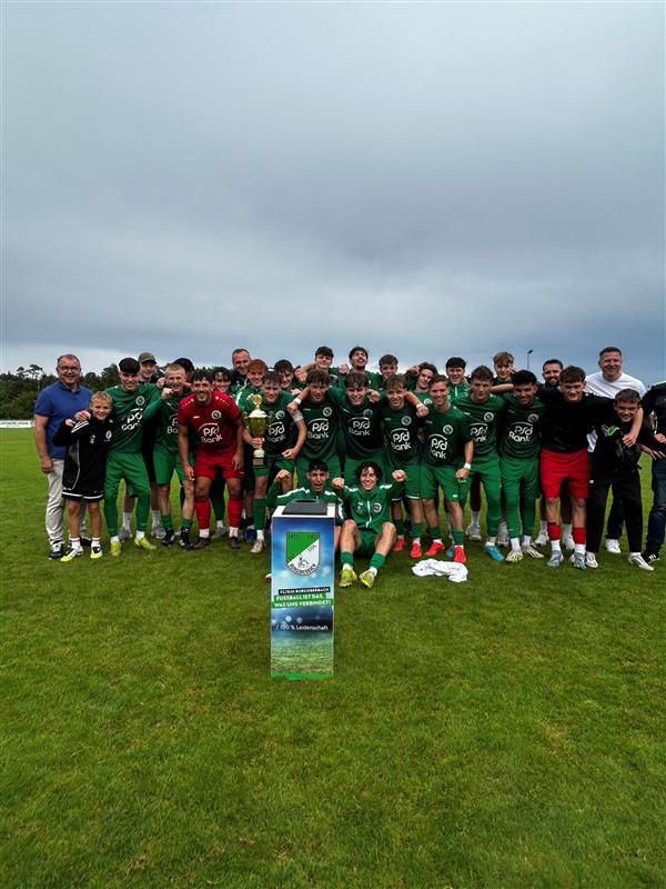Eine Fußballmannschaft in grünen Trikots jubelt gemeinsam auf dem Spielfeld mit einem großen Pokal und einer Turniertafel.