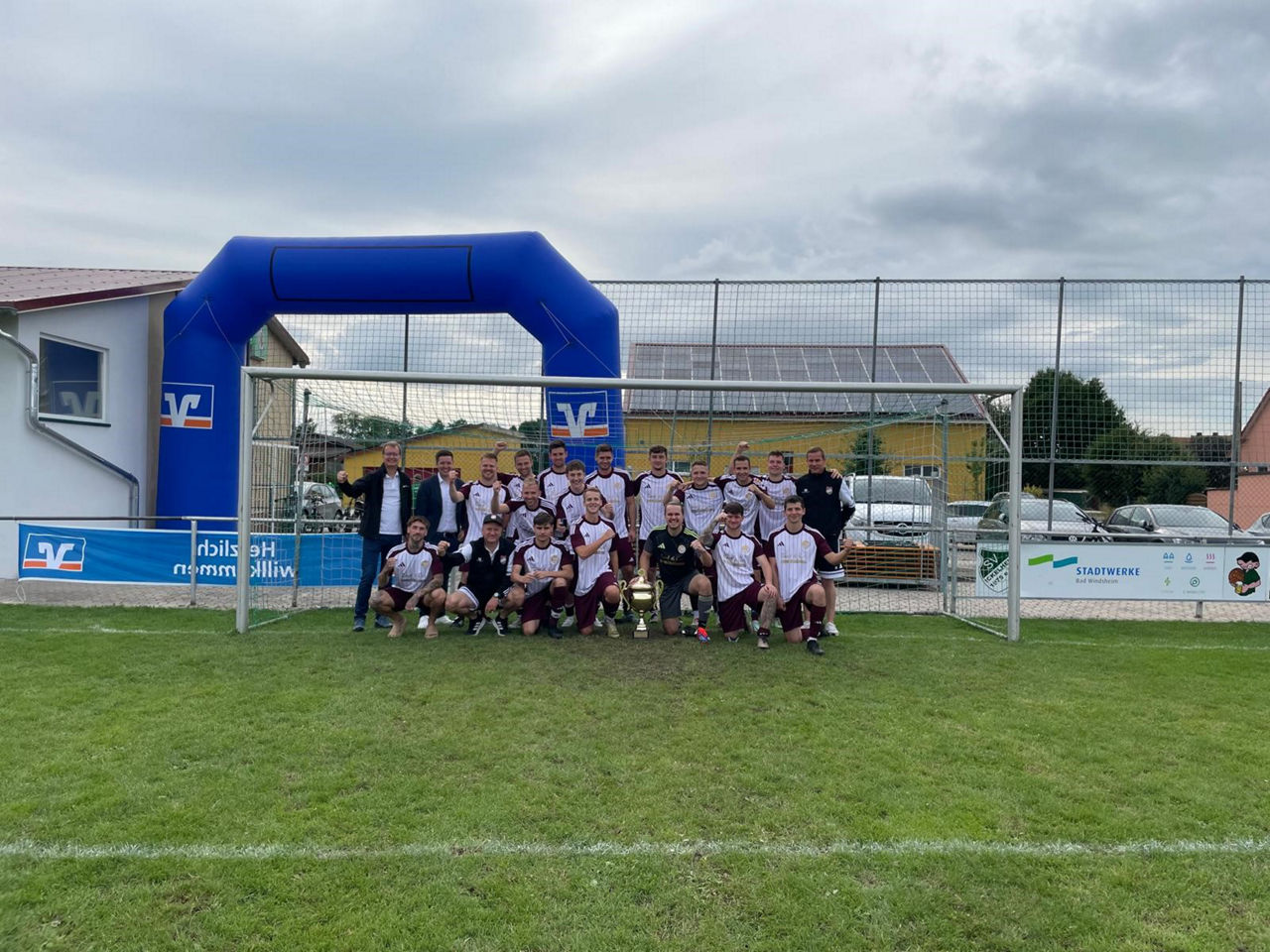 Eine Fußballmannschaft posiert jubelnd mit einem großen Pokal vor einem Tor auf dem Spielfeld.