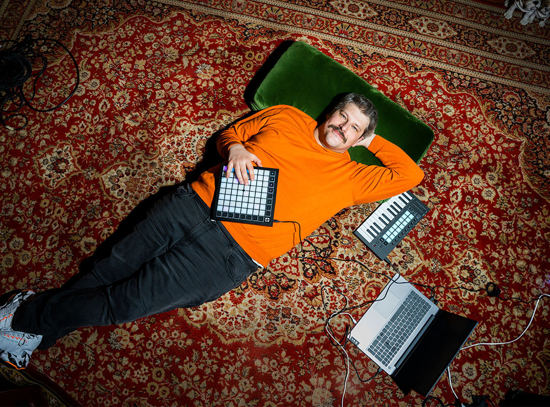 Ein Mann mit orangenfarbigen Pullover liegt entspannt auf dem Teppich