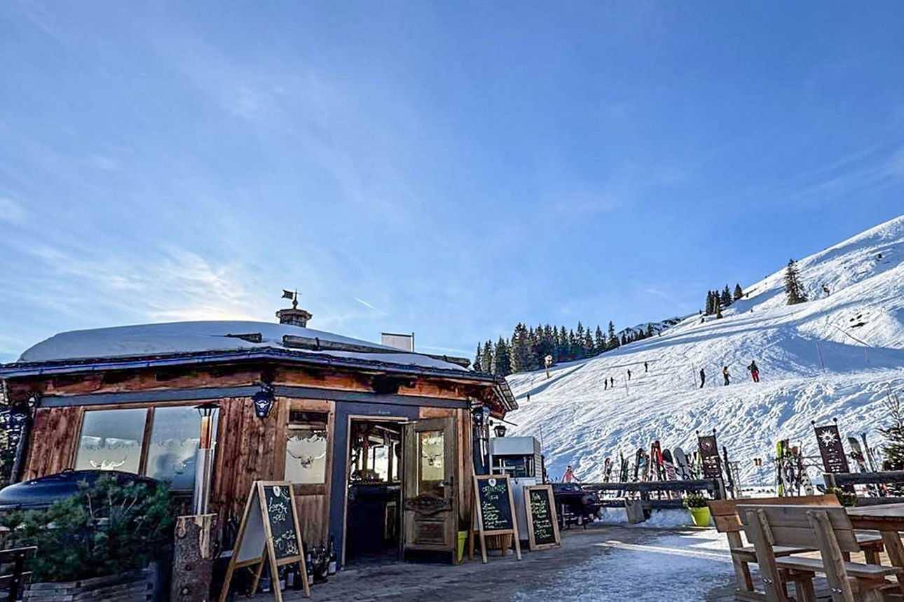 Eine Skihütte vor einem Berg mit Schnee. Es sind einige Ski-Fahrer zu sehen.