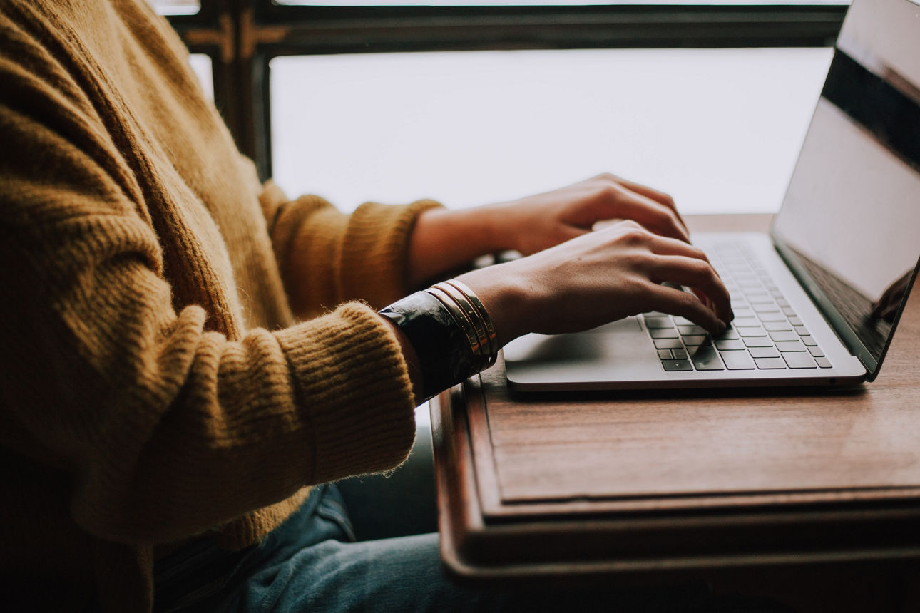 Frau sitzt neben einem Fenster an einem Holztisch und tippt auf einem Laptop