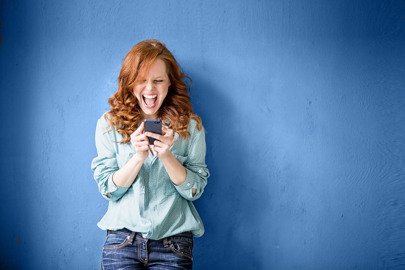 Lachende Frau, die auf ihr Handy schaut, steht vor blauer Wand