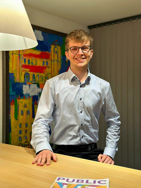 Junger Mann mit Brille steht vor einer Wand mit einem bunten Bild im Hintergrund und lächelt