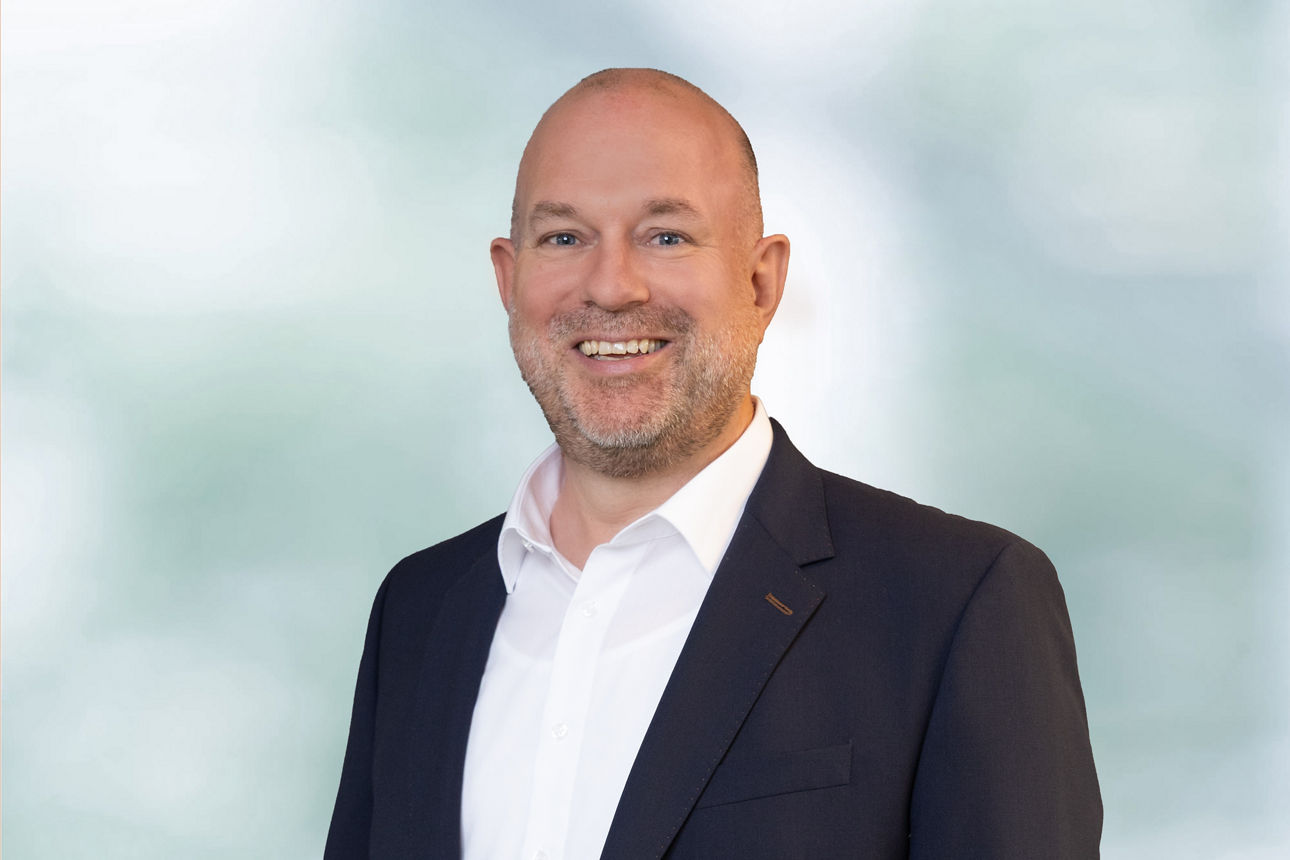 Portraitaufnahme unseres Kollegen Michael Soff. Er trägt ein weißes Hemd unter einem dunkelblauen Anzug. Michael Soff lächelt.
