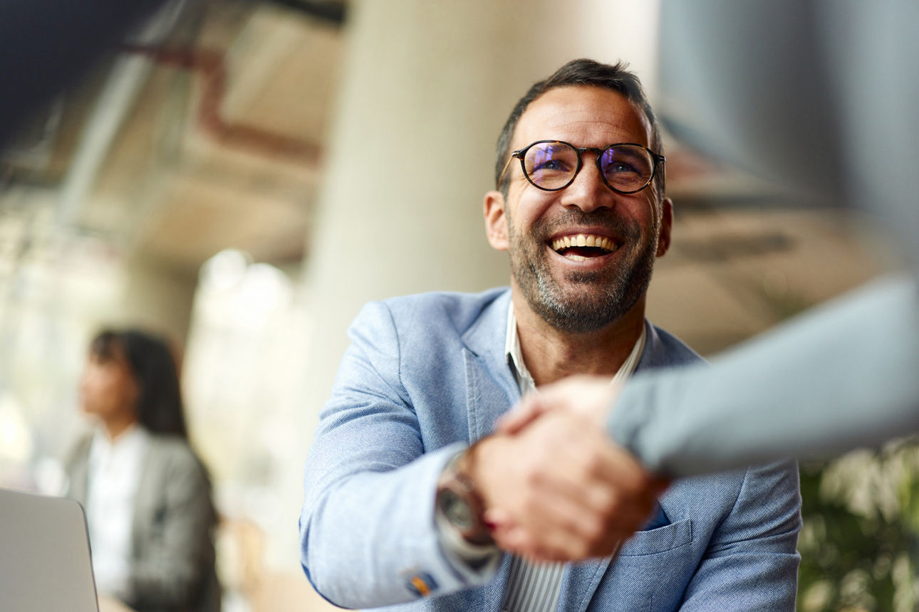 Ein lächelnder Mann mit Brille und blauem Sakko gibt einer anderen Person in einem modernen Büro die Hand.